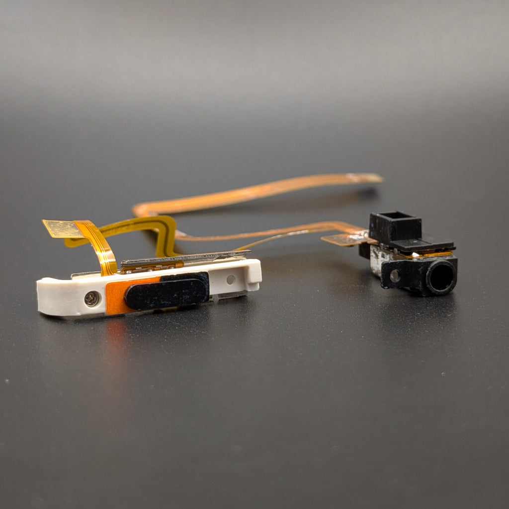A small electronic component assembly with a white and orange plastic housing, attached to a flexible copper ribbon cable, shown next to a separate black camera lens module on a dark gray background.