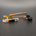 A small electronic component assembly with a white and orange plastic housing, attached to a flexible copper ribbon cable, shown next to a separate black camera lens module on a dark gray background.