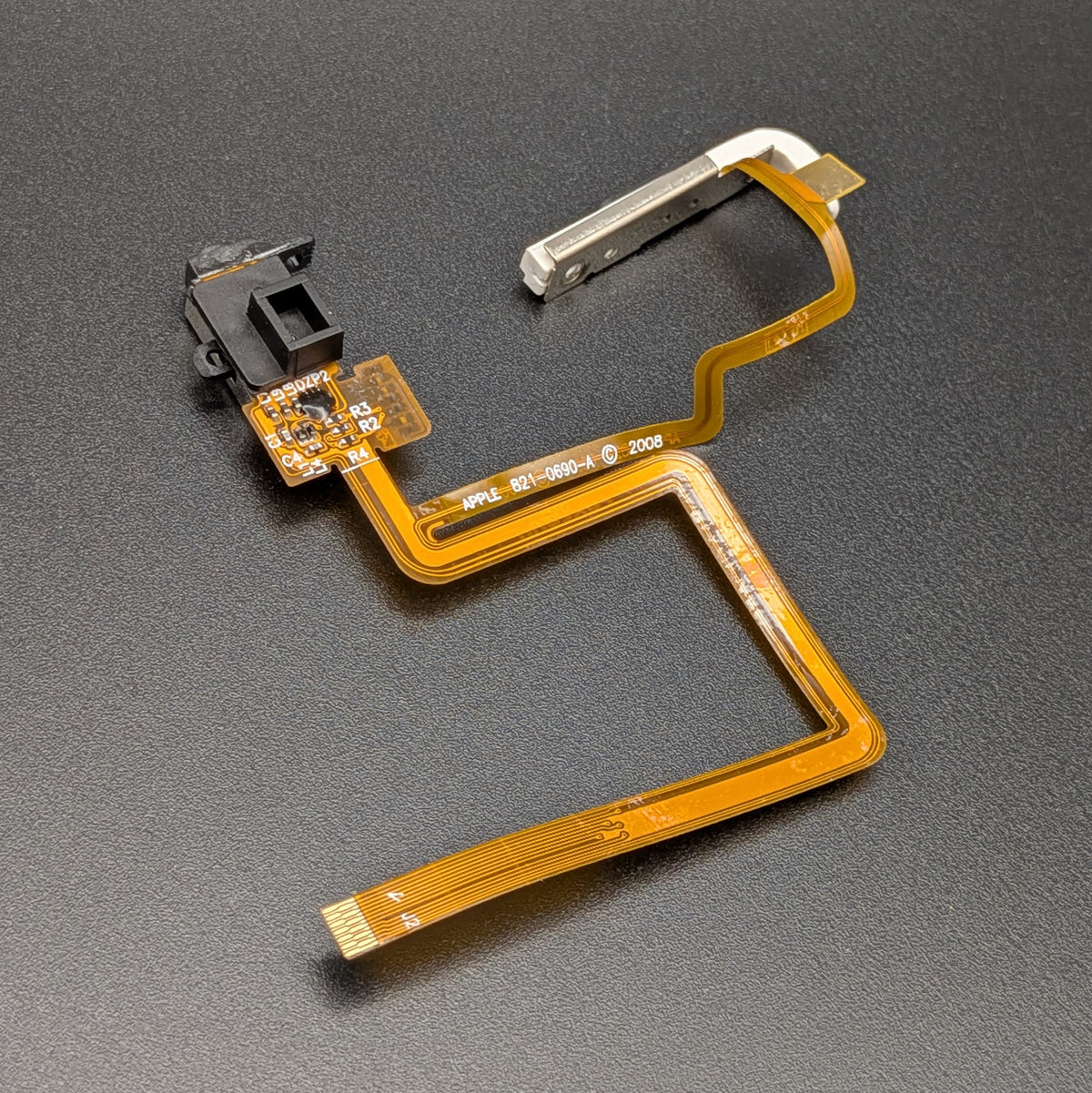 A replacement flex cable assembly for an iPod, featuring a gold-orange ribbon cable, a black plastic button housing, and a silver metal connector, shown on a dark textured background.