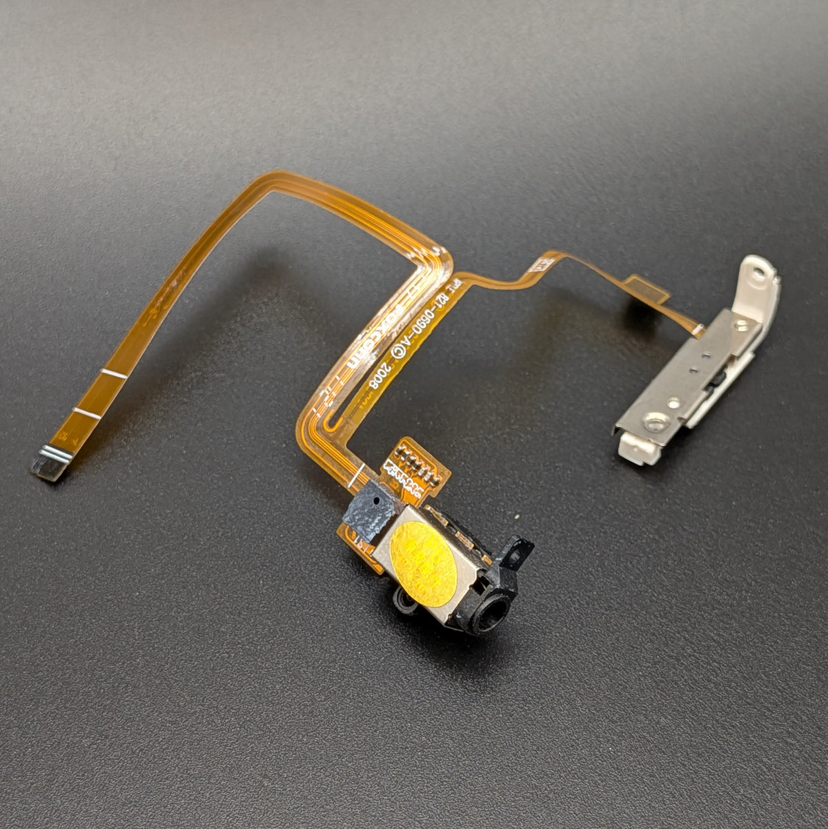 A replacement iPod flex cable assembly with a gold ribbon cable, an audio jack, and a silver metal connector, shown on a dark gray textured background.