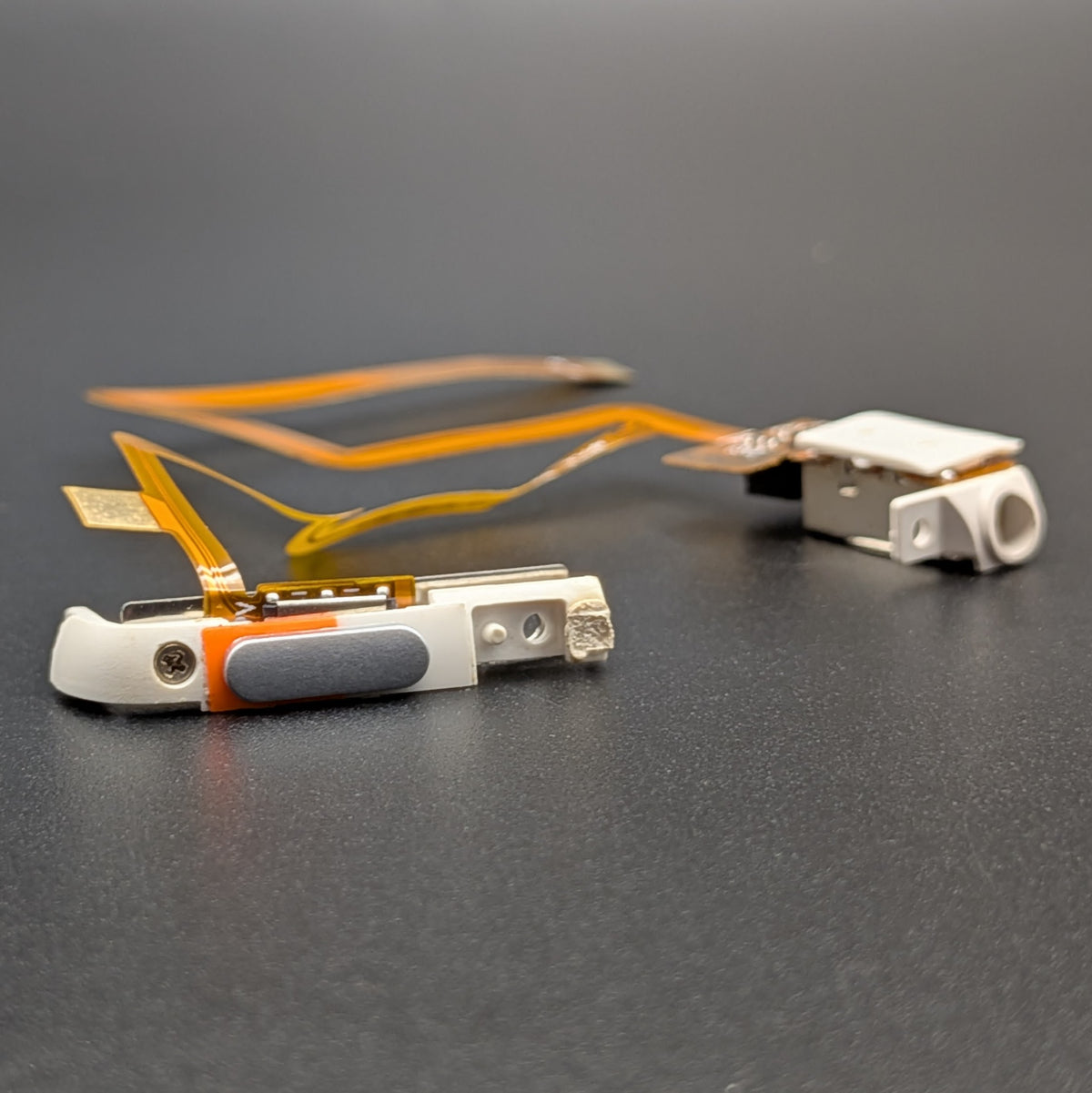 A close-up of a small electronic repair part or replacement component on a dark gray surface, featuring a white plastic body, orange accents, a gray grip, and flexible orange circuitry leading to another small white and silver component.