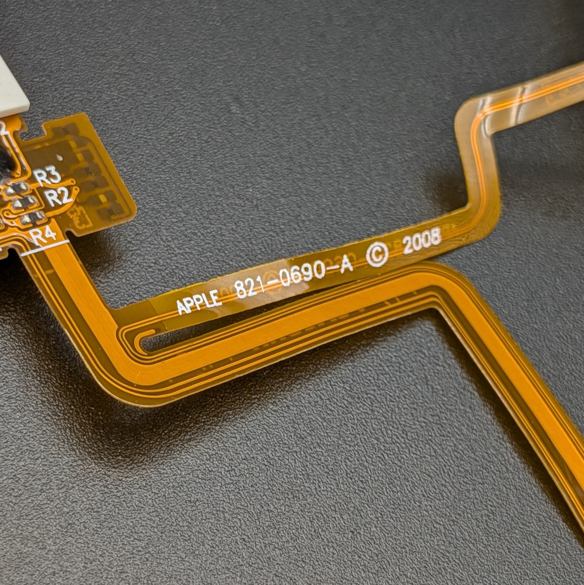 A close-up of a flexible circuitry ribbon cable with gold traces and white printed text including 'APPLE 821-0690-A' and '2008', laid on a dark textured surface.