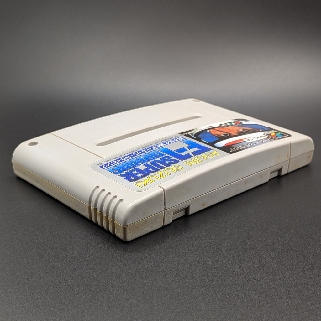 Aguri Suzuki F-1 Super Driving video game cartridge for Nintendo's Super Famicom (SFC) Entertainment System, shown laying on its back, facing upwards showing the front label, at a 3/4 angle on a dark grey background.
