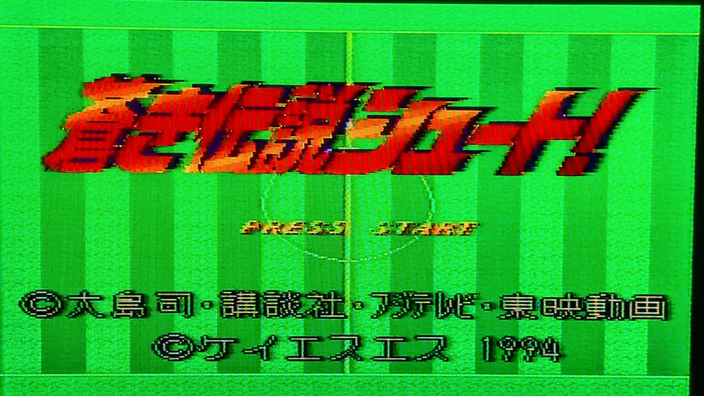 The video game title screen for Aoki Densetsu Shoot for Nintendo's Super Famicom (SFC), featuring large red and black stylised Japanese text over a green striped football field in the background, plus 'PRESS START' and copyright text for 1994. This captures the game in working order.