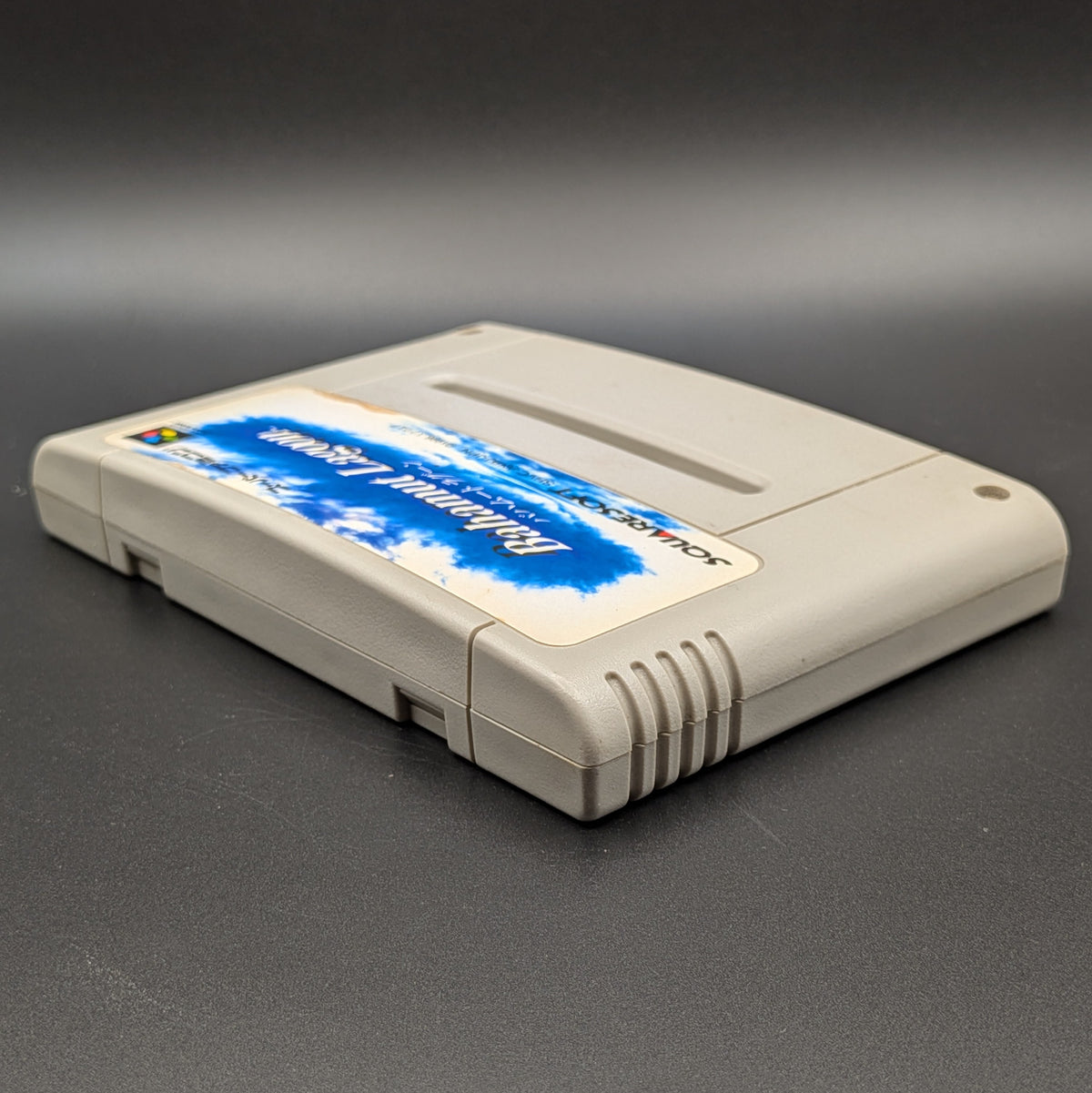 Bahamut Lagoon video game cartridge for Nintendo's Super Famicom (SFC) Entertainment System, shown laying on its back with the label facing upwards. Showing the left hand side and top edge. It sits on a dark grey background.