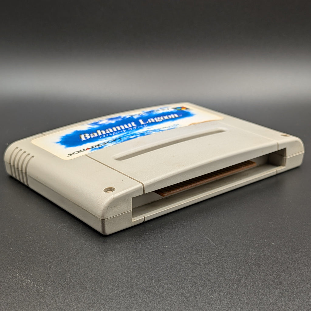 Bahamut Lagoon video game cartridge for Nintendo's Super Famicom (SFC) Entertainment System, shown laying on its back with the label facing upwards. Showing the left hand side and bottom slot. It sits on a dark grey background