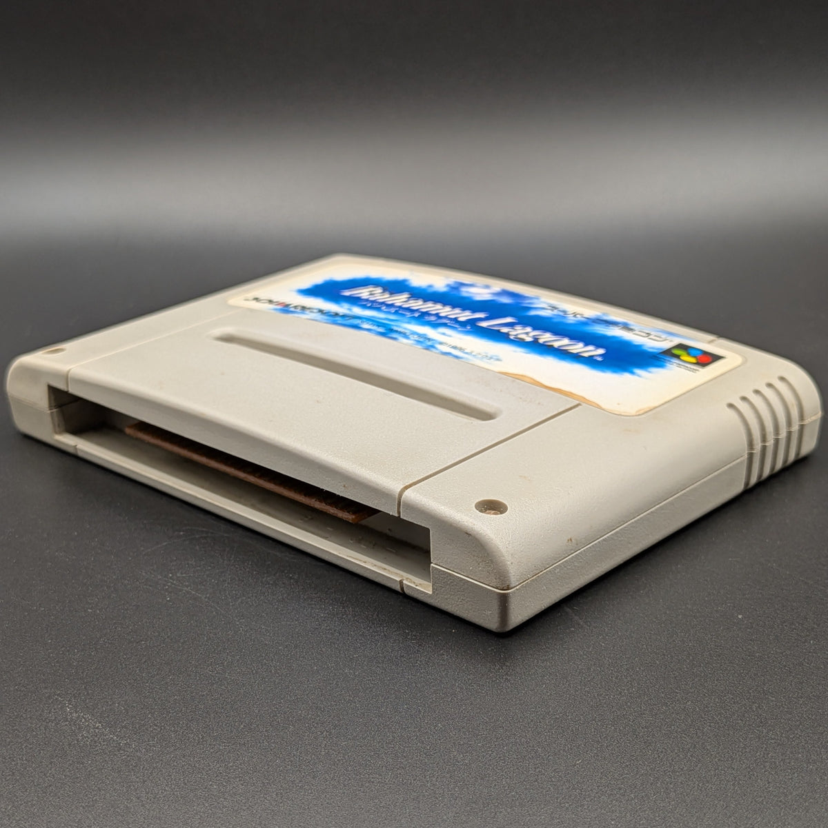 Bahamut Lagoon video game cartridge for Nintendo's Super Famicom (SFC) Entertainment System, shown laying on its back with the label facing upwards. Showing the right hand side and bottom slot. It sits on a dark grey background