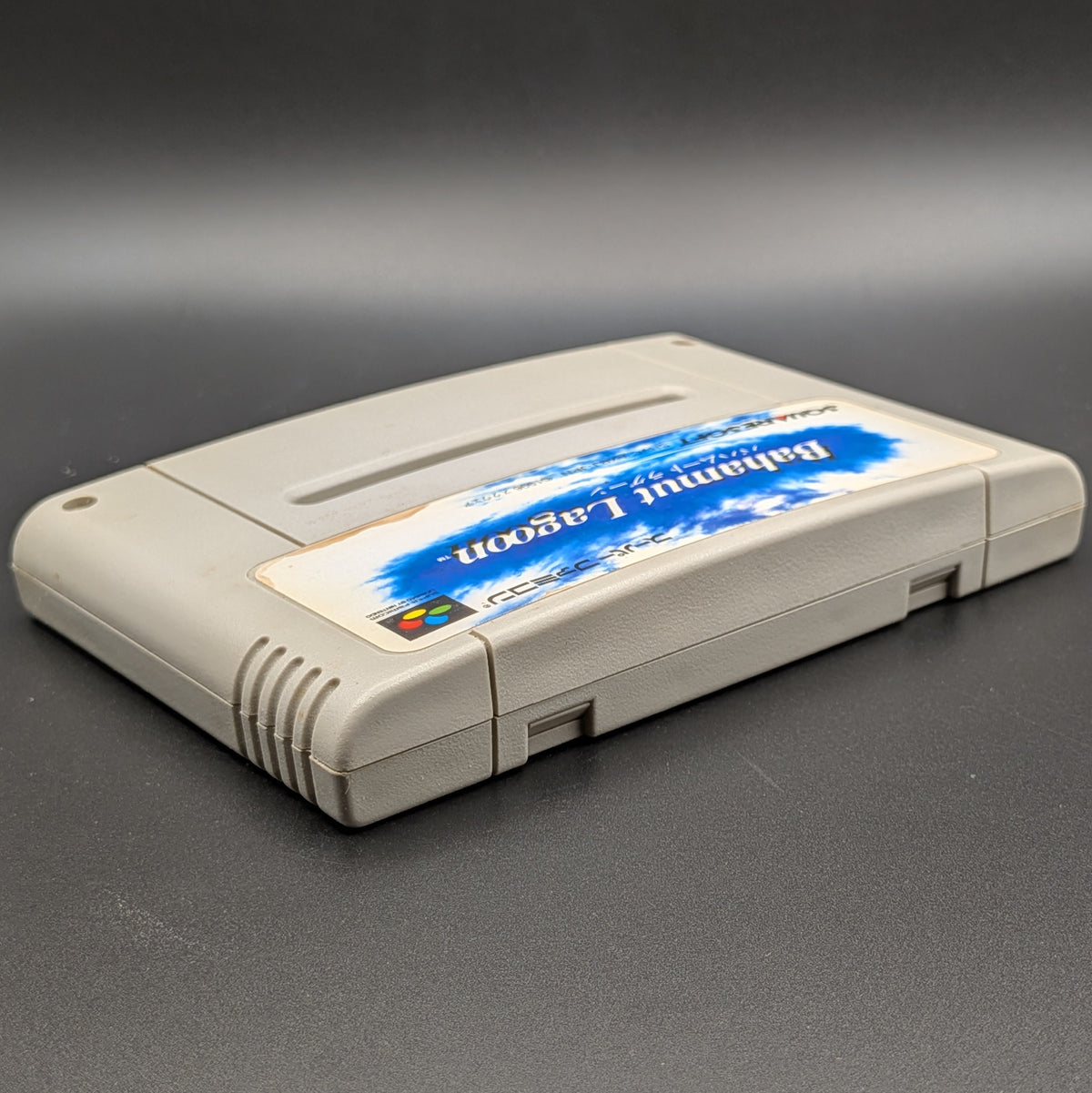 Bahamut Lagoon video game cartridge for Nintendo's Super Famicom (SFC) Entertainment System, shown laying on its back with the label facing upwards. Showing the right hand side and top edge. It sits on a dark grey background.