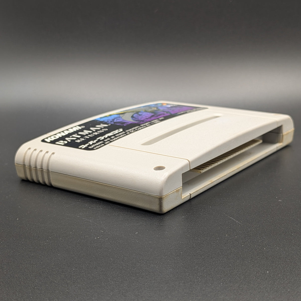 Batman Returns video game cartridge for Nintendo's Super Famicom (SFC) Entertainment System, shown laying on its back with the label facing upwards. Showing the left hand side and bottom slot. It sits on a dark grey background.