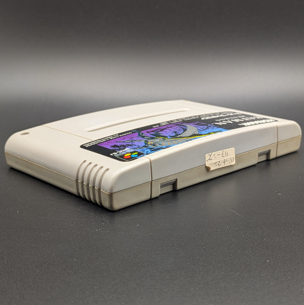 Batman Returns video game cartridge for Nintendo's Super Famicom (SFC) Entertainment System, shown laying on its back with the label facing upwards. Showing the right hand side and top edge. It sits on a dark grey background. On the top edge is a small white sticker with Japanese hand writing on it.