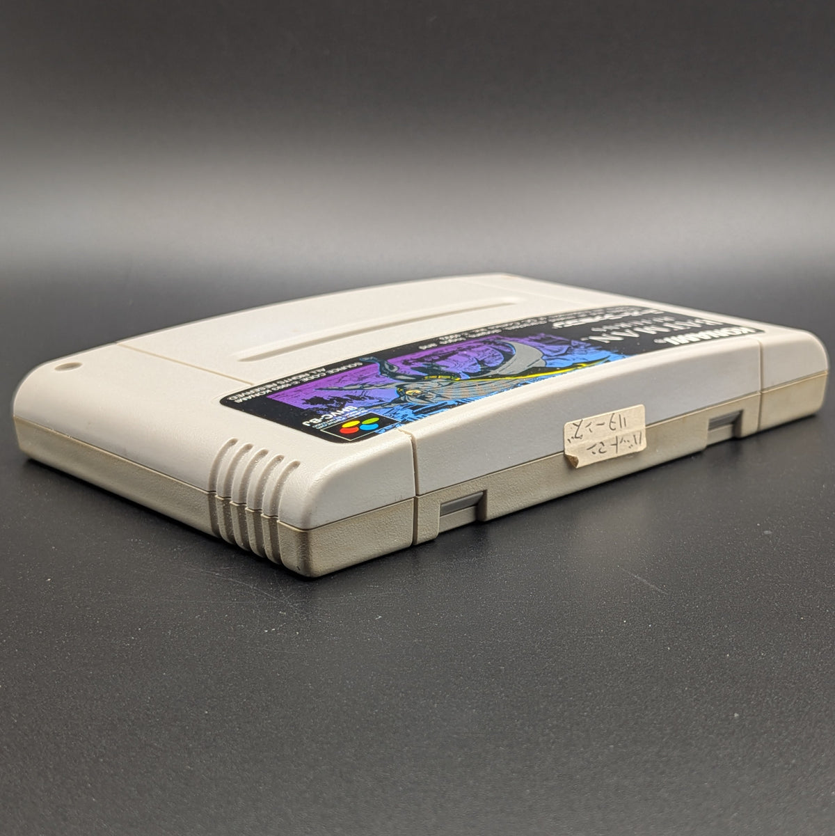 Batman Returns video game cartridge for Nintendo's Super Famicom (SFC) Entertainment System, shown laying on its back with the label facing upwards. Showing the right hand side and top edge. It sits on a dark grey background. On the top edge is a small white sticker with Japanese hand writing on it.