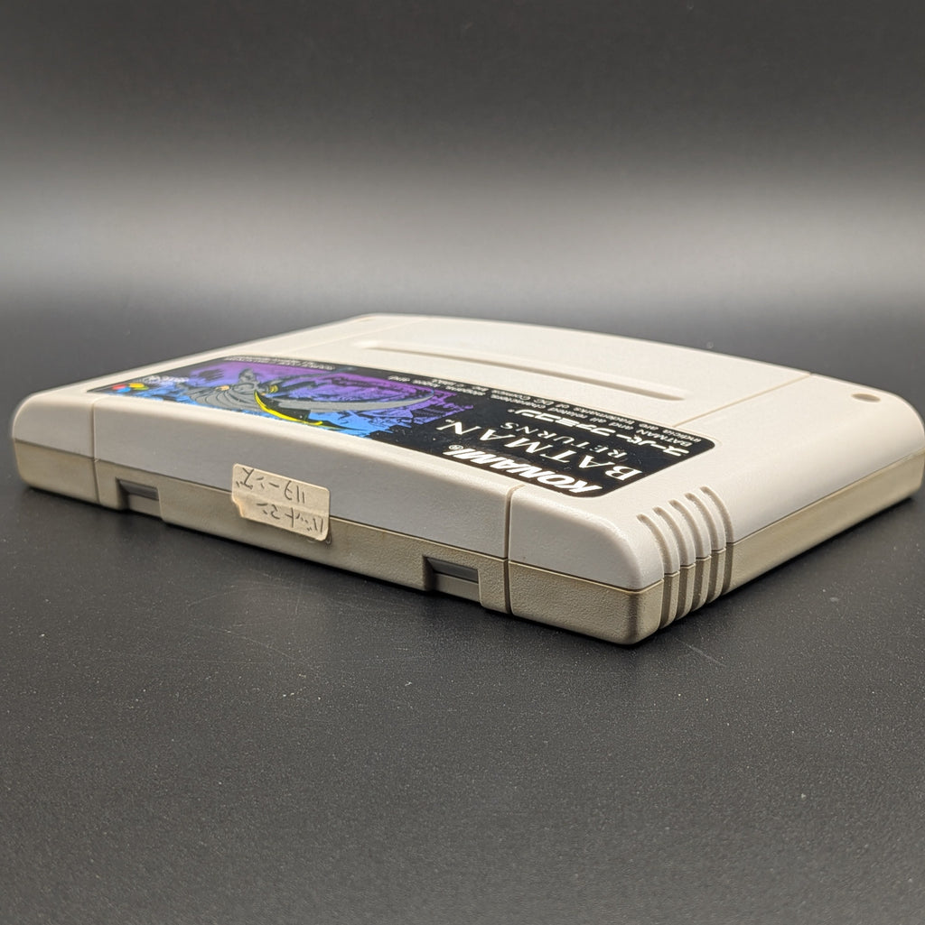 Batman Returns video game cartridge for Nintendo's Super Famicom (SFC) Entertainment System, shown laying on its back with the label facing upwards. Showing the left hand side and top edge. It sits on a dark grey background. On the top edge is a small white sticker with Japanese hand writing on it.