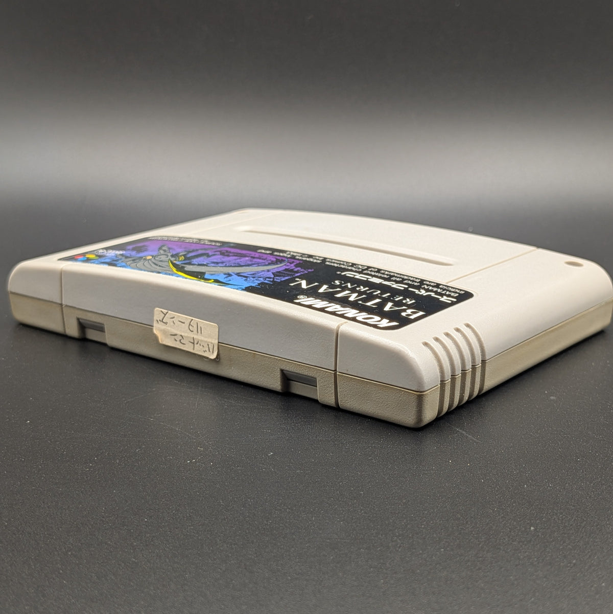 Batman Returns video game cartridge for Nintendo's Super Famicom (SFC) Entertainment System, shown laying on its back with the label facing upwards. Showing the left hand side and top edge. It sits on a dark grey background. On the top edge is a small white sticker with Japanese hand writing on it.