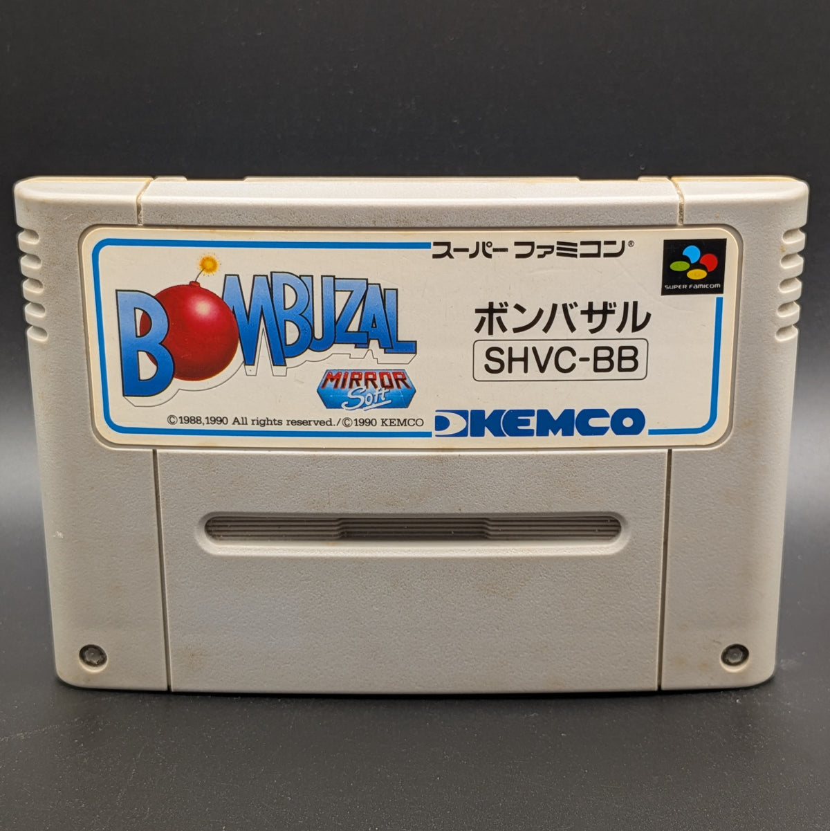 A close up of Bombuzal video game cartridge for Nintendo's Super Famicom (SFC) Entertainment System, shown standing up and facing forward on a dark grey background.