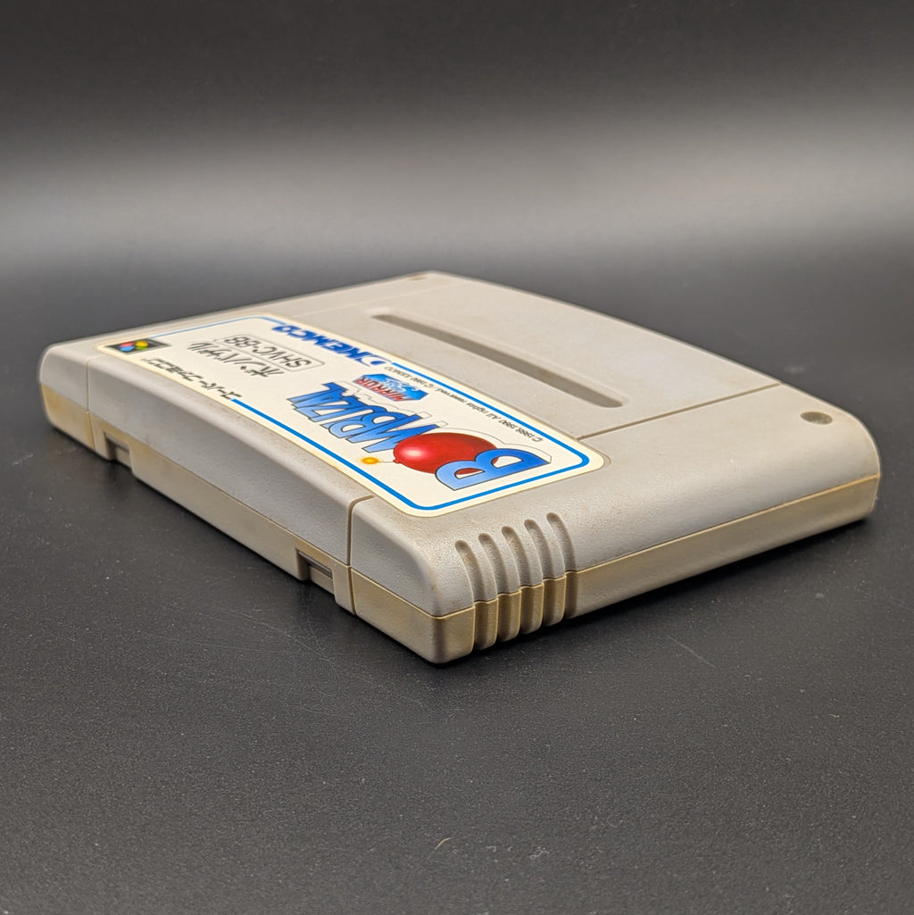 Bombuzal video game cartridge for Nintendo's Super Famicom (SFC) Entertainment System, shown laying on its back with the label facing upwards. Showing the left hand side and top edge. It sits on a dark grey background. 