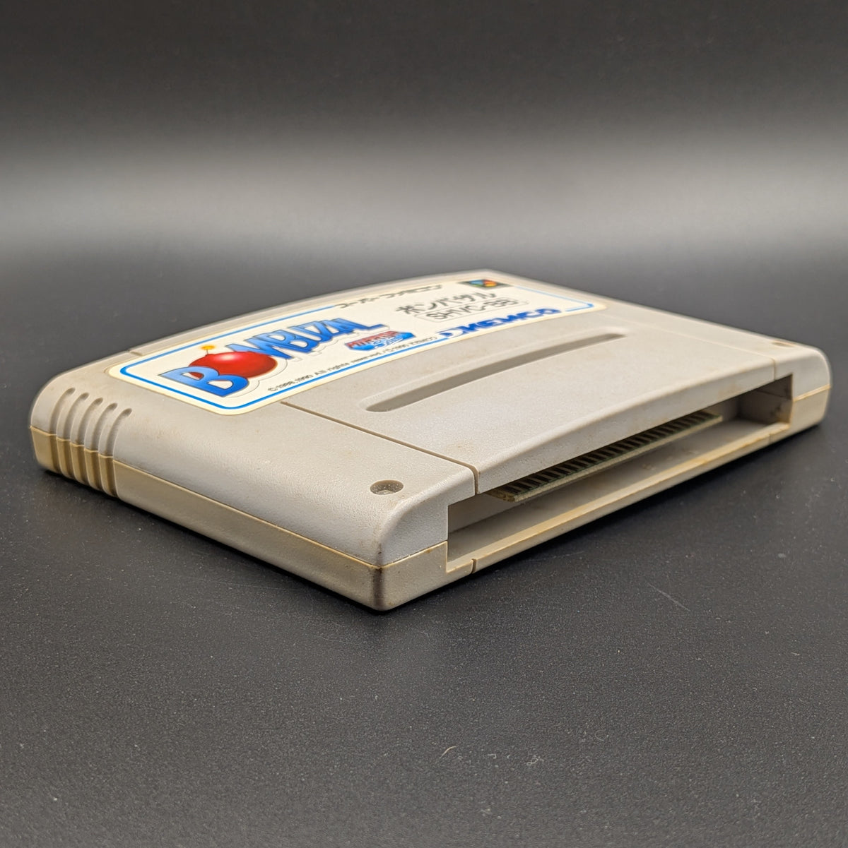 Bombuzal video game cartridge for Nintendo's Super Famicom (SFC) Entertainment System, shown laying on its back with the label facing upwards. Showing the left hand side and bottom slot. It sits on a dark grey background. 