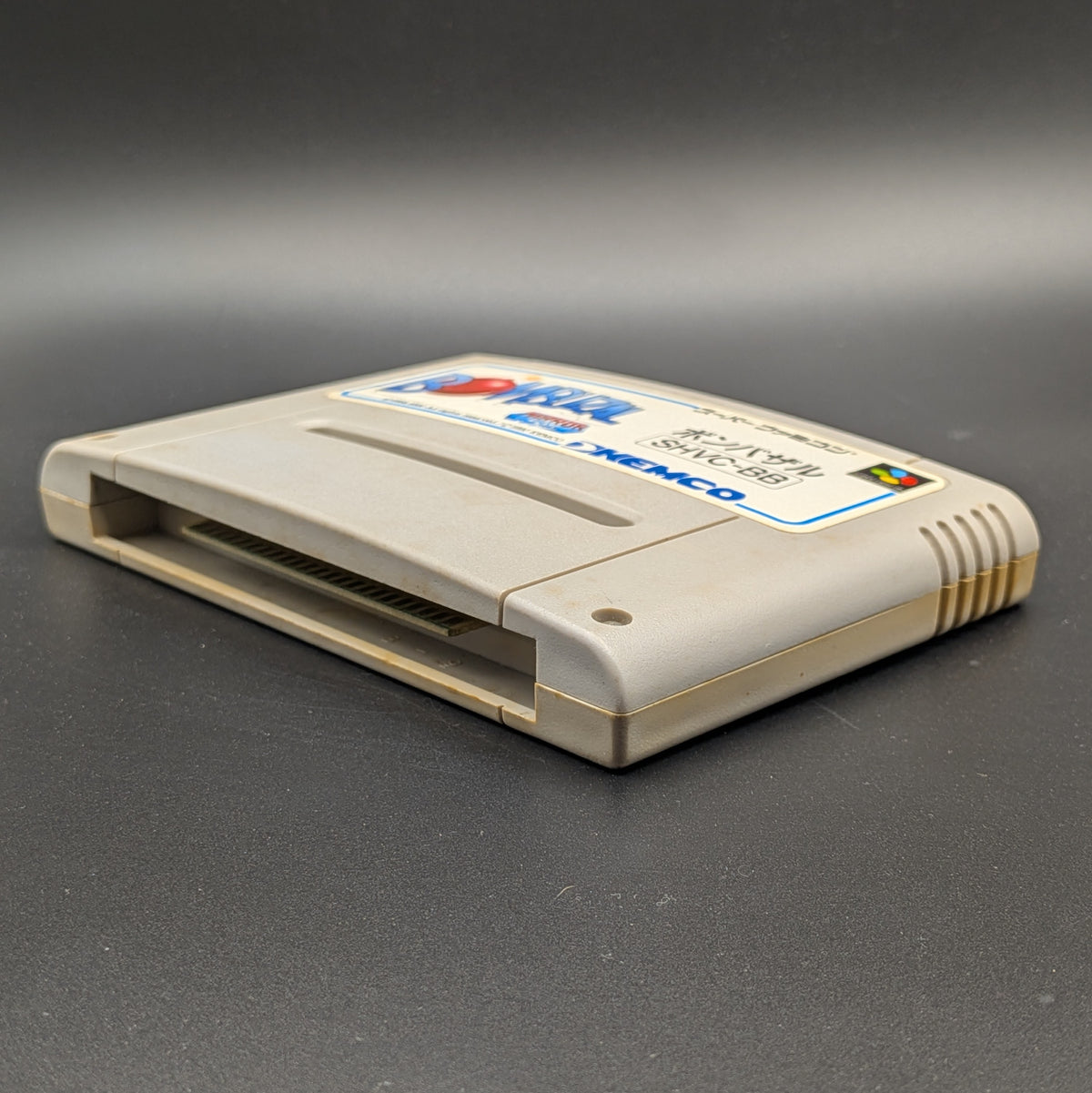 Bombuzal video game cartridge for Nintendo's Super Famicom (SFC) Entertainment System, shown laying on its back with the label facing upwards. Showing the right hand side and bottom slot. It sits on a dark grey background. 
