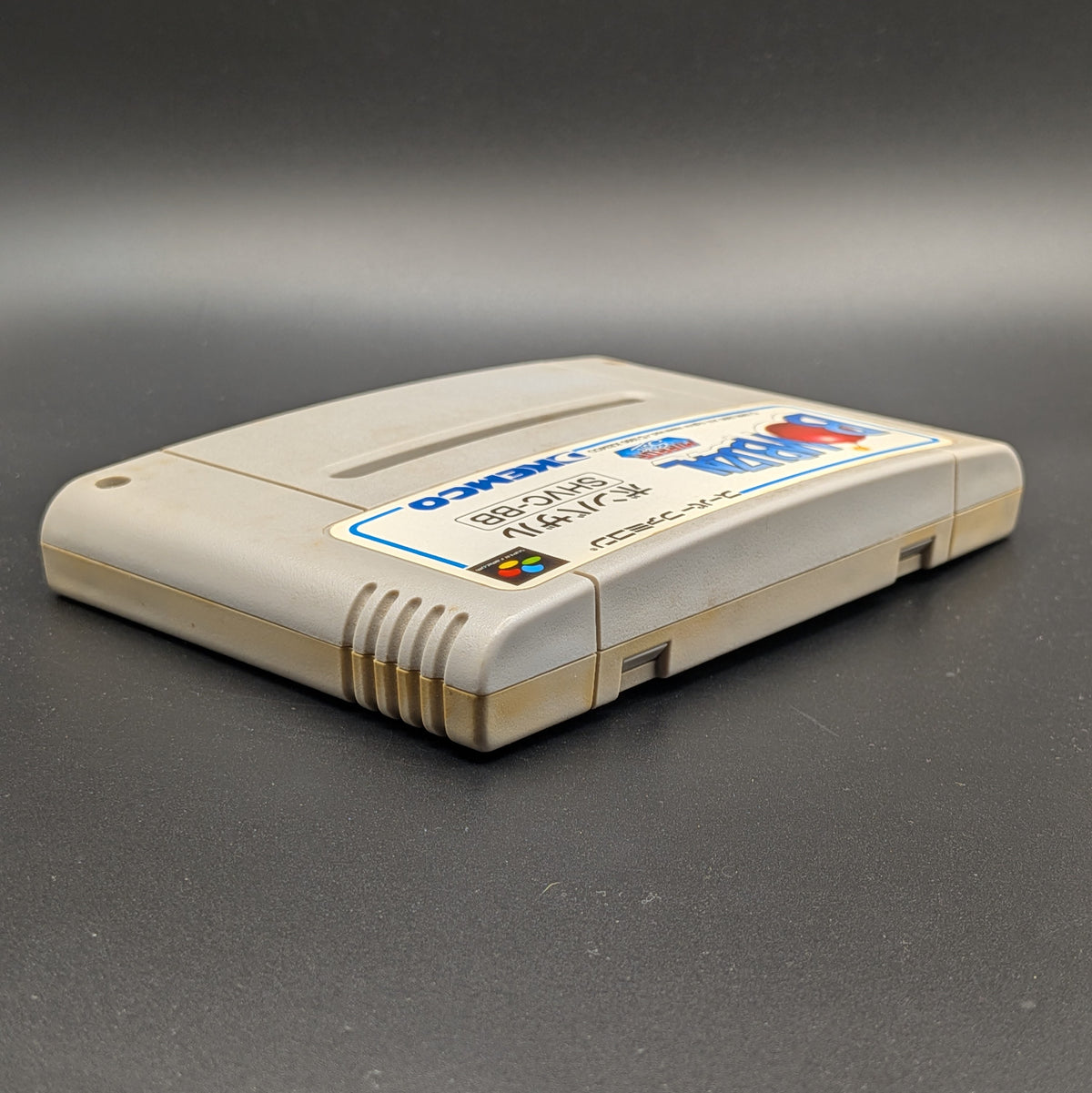 Bombuzal video game cartridge for Nintendo's Super Famicom (SFC) Entertainment System, shown laying on its back with the label facing upwards. Showing the right hand side and top edge. It sits on a dark grey background. 