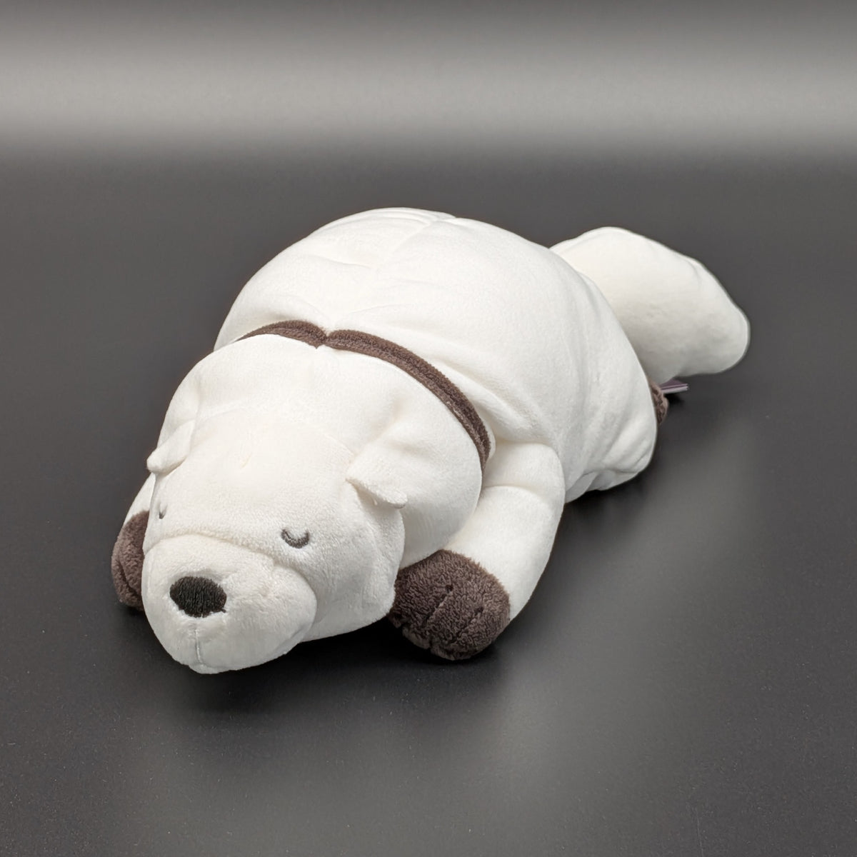 A plush Bond Forger dog toy shown lying on its belly, facing forwards at 3/4 view angle. Sitting on a dark grey surface, with soft white fur and brownish grey accents on its paws, snout, and collar. From the anime/manga series Spy X Family, made by Club Mochi-Mochi.