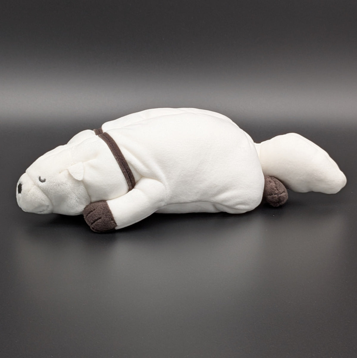 A plush Bond Forger dog toy shown lying on its belly, facing side on. Sitting on a dark grey surface, with soft white fur and brownish grey accents on its paws, snout, and collar. From the anime/manga series Spy X Family, made by Club Mochi-Mochi.