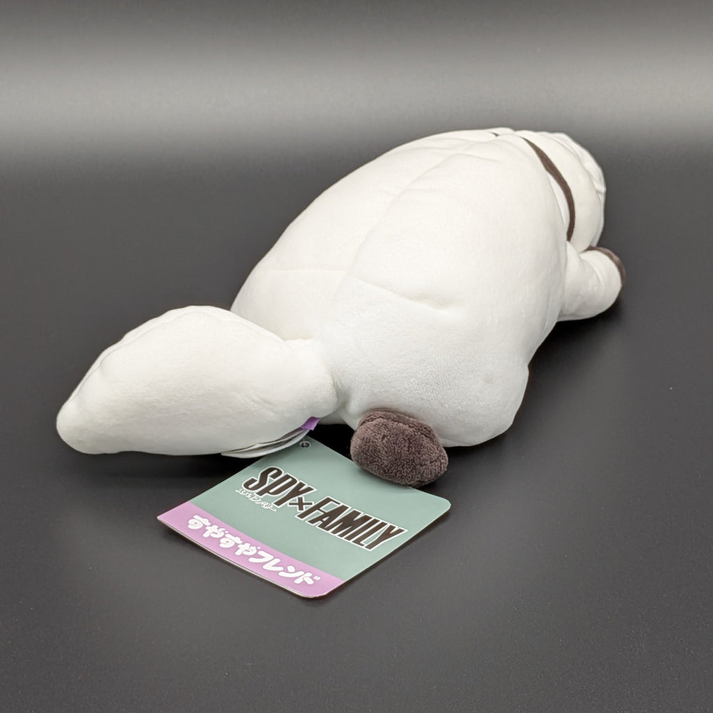 A plush Bond Forger dog toy shown lying on its belly, facing backwards at 3/4 view angle also showing the official Spy X Faimly tag. Sitting on a dark grey surface, with soft white fur and brownish grey accents on its paws, snout, and collar. From the anime/manga series Spy X Family, made by Club Mochi-Mochi.