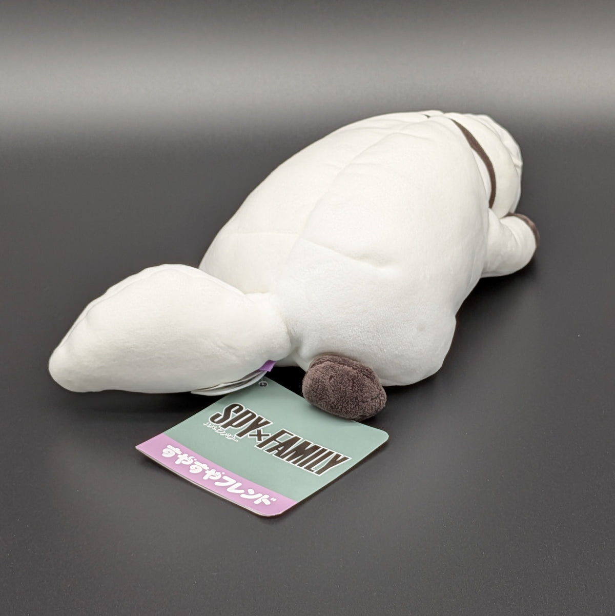 A plush Bond Forger dog toy shown lying on its belly, facing backwards at 3/4 view angle also showing the official Spy X Faimly tag. Sitting on a dark grey surface, with soft white fur and brownish grey accents on its paws, snout, and collar. From the anime/manga series Spy X Family, made by Club Mochi-Mochi.