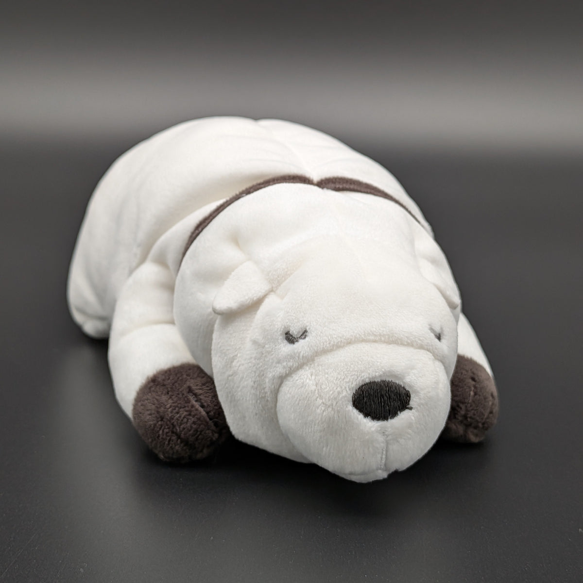 A plush Bond Forger dog toy shown lying on its belly, facing forwards at 3/4 view angle. Sitting on a dark grey surface, with soft white fur and brownish grey accents on its paws, snout, and collar. From the anime/manga series Spy X Family, made by Club Mochi-Mochi.