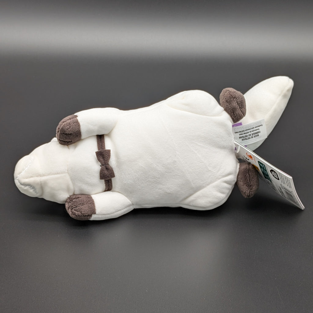 A plush Bond Forger dog toy shown lying on its side to reveal his belly, you can see his bow tie. Sitting on a dark grey surface, with soft white fur and brownish grey accents on its paws, snout, and collar. From the anime/manga series Spy X Family, made by Club Mochi-Mochi.