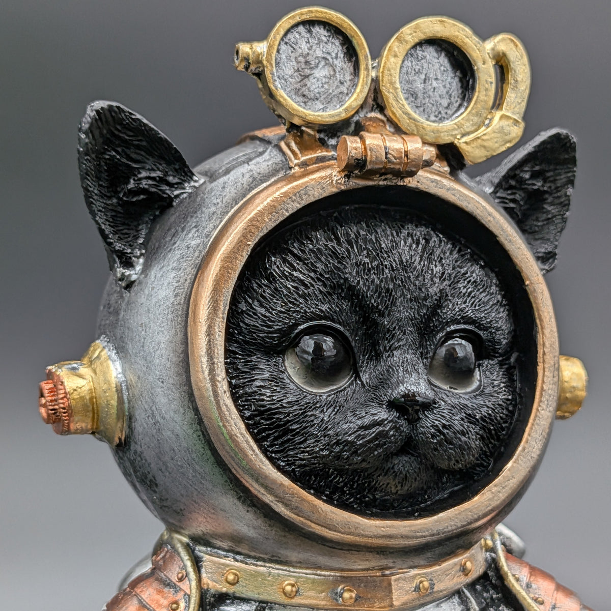 A close-up of a decorative cat figurine wearing a vintage space helmet with brass-toned goggles and metallic details, shown against a soft gray background.