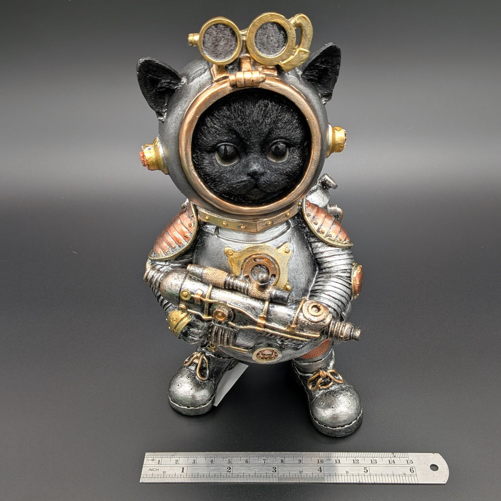 A decorative figurine of a black cat dressed in a detailed steampunk-style space suit, standing on a dark gray background with a metal ruler at the bottom for scale.