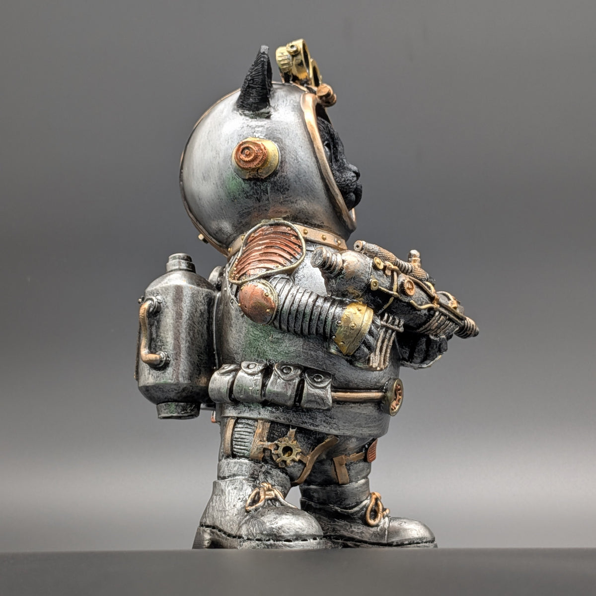 A small decorative figurine of a stylized cat dressed in a detailed, weathered steampunk-style space suit. The suit features rusted copper panels, brass-toned metal accents, and a silver-gray body, set against a neutral gray studio background.