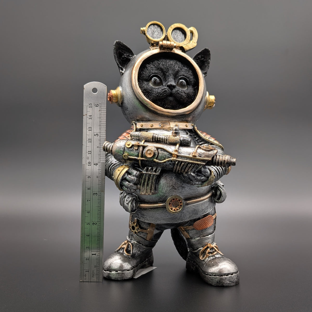 A steampunk-style figurine of a black cat wearing a detailed silver and gold space suit, holding a futuristic gun, standing next to a metal ruler on a dark gray background.
