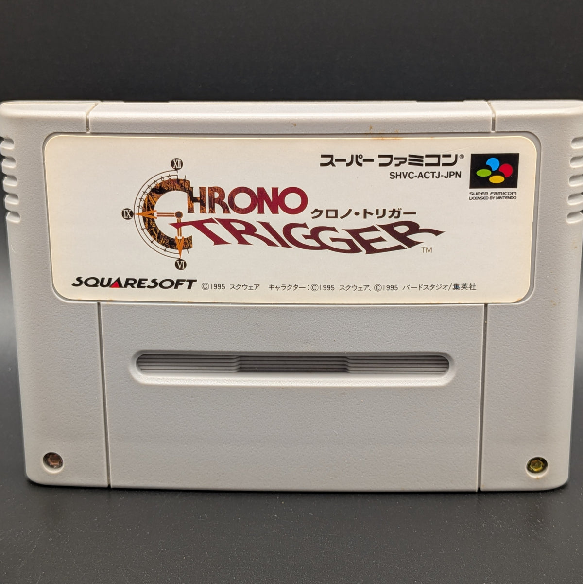 A close up of Chrono Trigger video game cartridge for Nintendo's Super Famicom (SFC) Entertainment System, shown standing up and facing forward on a dark grey background.
