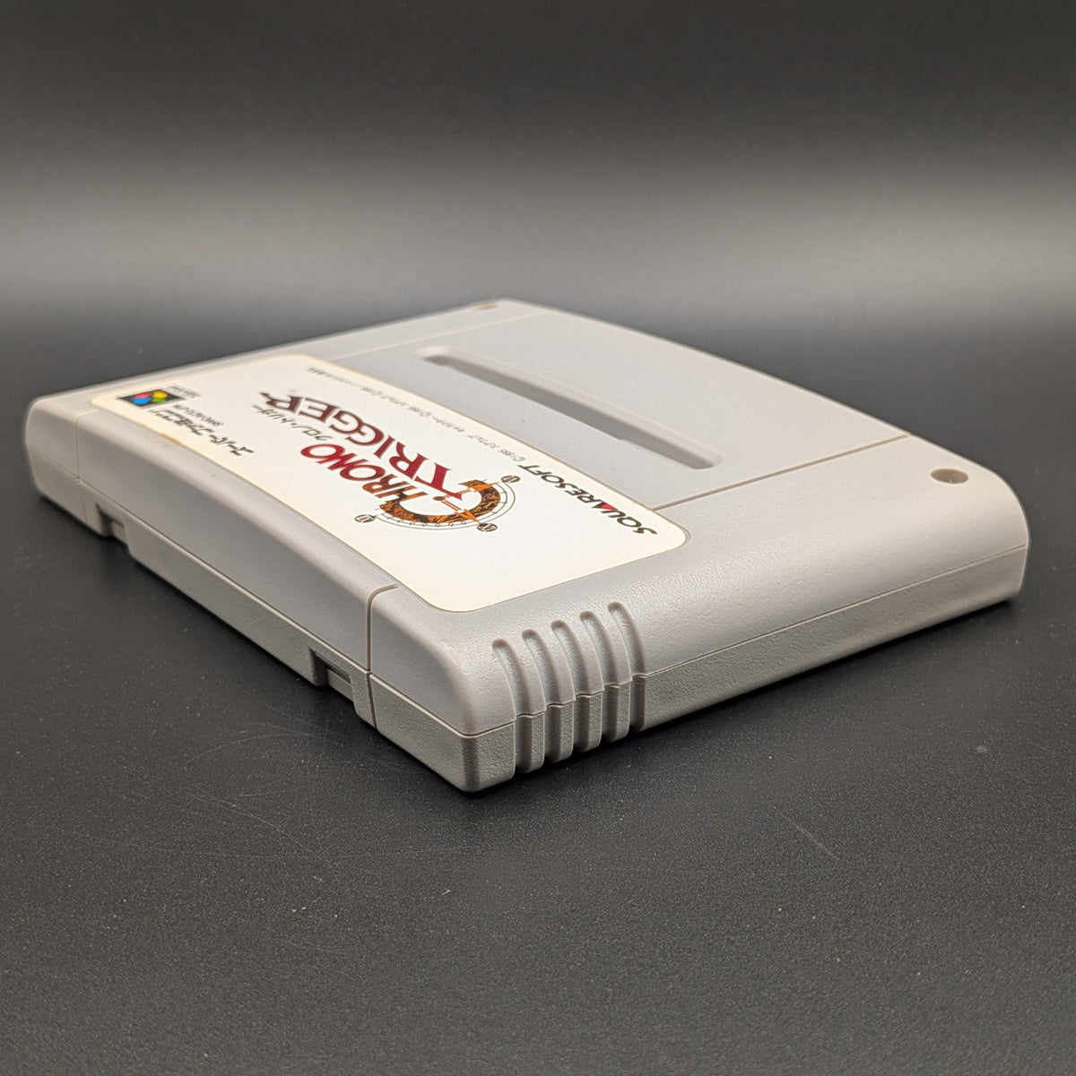 Chrono Trigger video game cartridge for Nintendo's Super Famicom (SFC) Entertainment System, shown laying on its back with the label facing upwards. Showing the left hand side and top edge. It sits on a dark grey background.