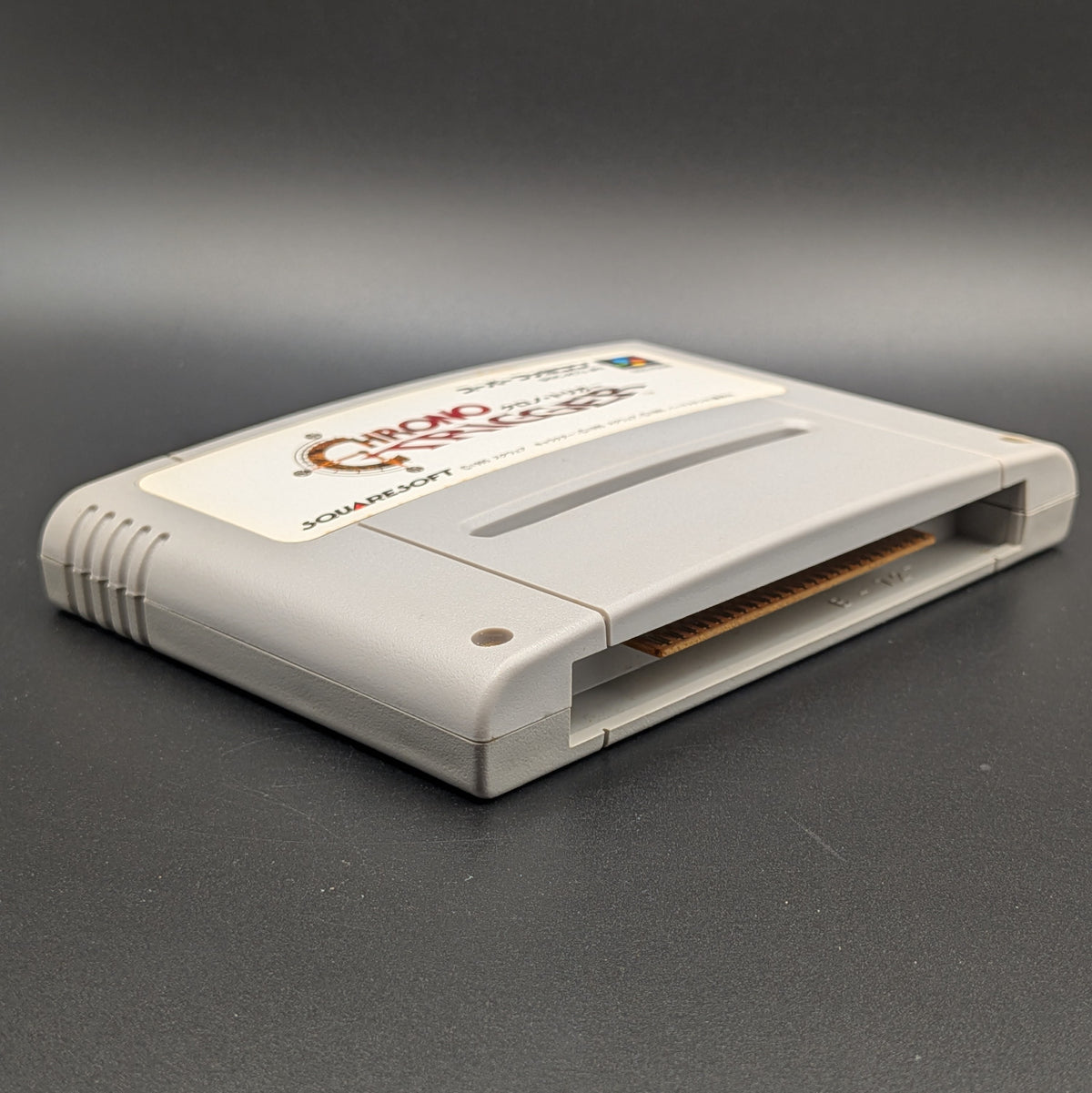 Chrono Trigger video game cartridge for Nintendo's Super Famicom (SFC) Entertainment System, shown laying on its back with the label facing upwards. Showing the left hand side and bottom slot. It sits on a dark grey background. 