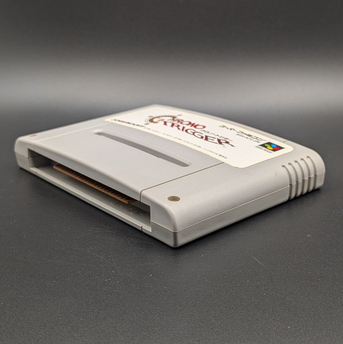 Chrono Trigger video game cartridge for Nintendo's Super Famicom (SFC) Entertainment System, shown laying on its back with the label facing upwards. Showing the right hand side and bottom slot. It sits on a dark grey background.
