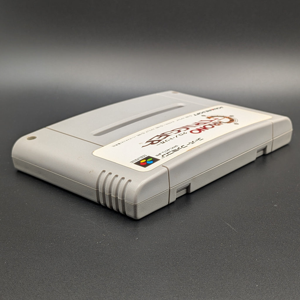 Chrono Trigger video game cartridge for Nintendo's Super Famicom (SFC) Entertainment System, shown laying on its back with the label facing upwards. Showing the right hand side and top edge. It sits on a dark grey background. 