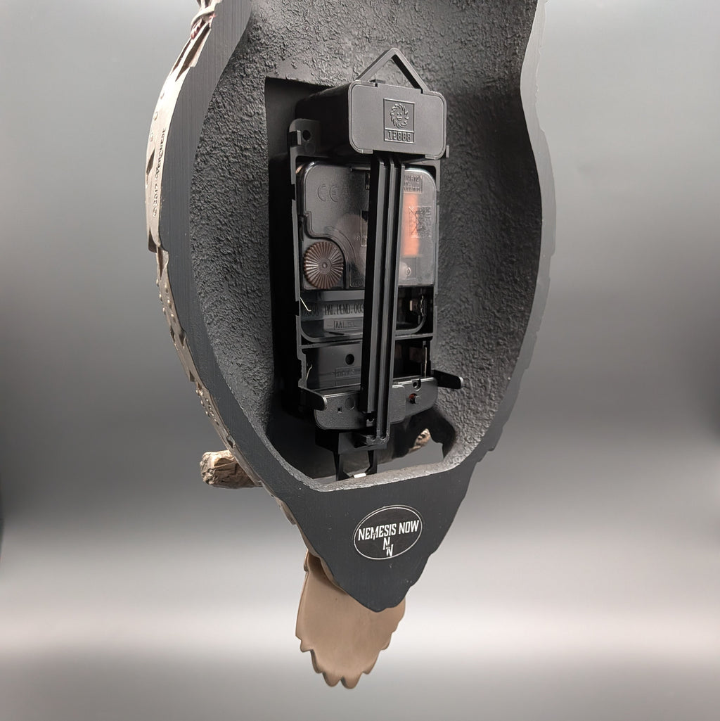 The back of a clock in a bird-shaped housing with the tail of the bird as the pendulum attachment at the bottom. The housing has a visible circular logo reading 'NEMESIS NOW'.