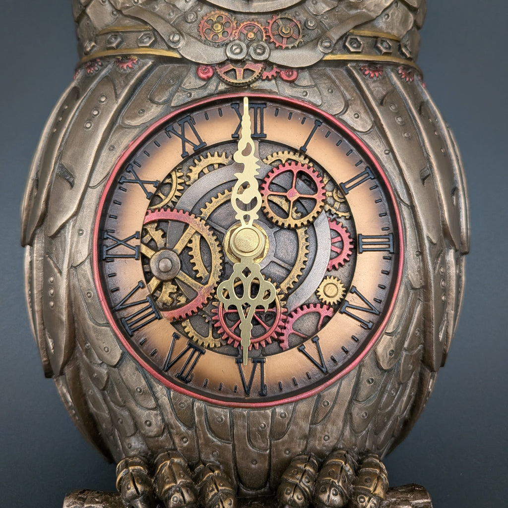 A close-up of an ornate steampunk owl clock with a detailed body, Roman numerals, and visible internal gears in gold, red, and silver tones against a dark gray background.