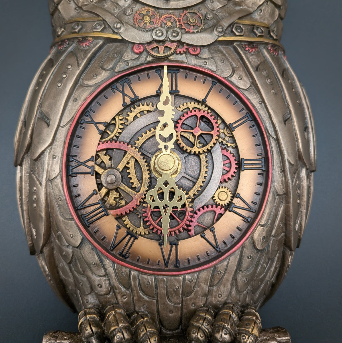 A close-up of an ornate steampunk owl clock with a detailed body, Roman numerals, and visible internal gears in gold, red, and silver tones against a dark gray background.