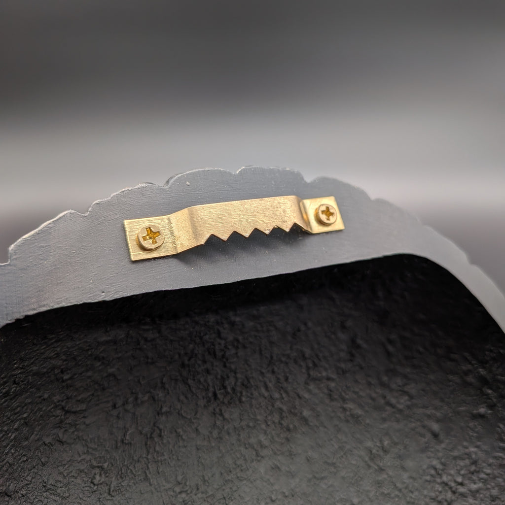Close-up of a wall hanging bracket, featuring a small gold metal bracket with two screws on the curved top edge, set against a dark gray gradient background.