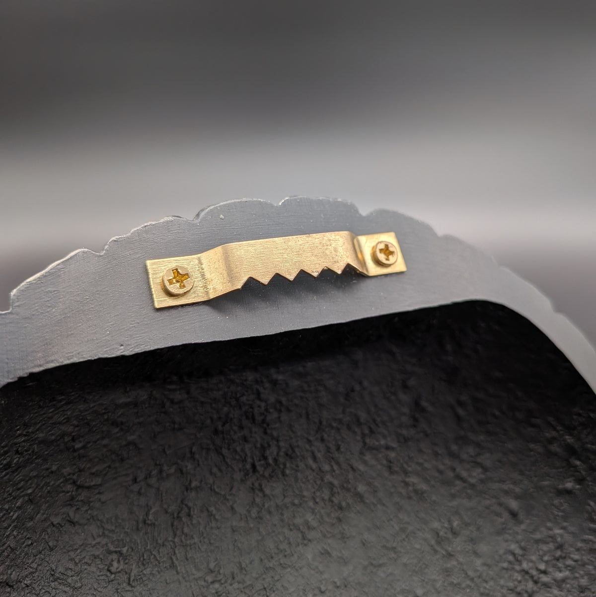 Close-up of a wall hanging bracket, featuring a small gold metal bracket with two screws on the curved top edge, set against a dark gray gradient background.