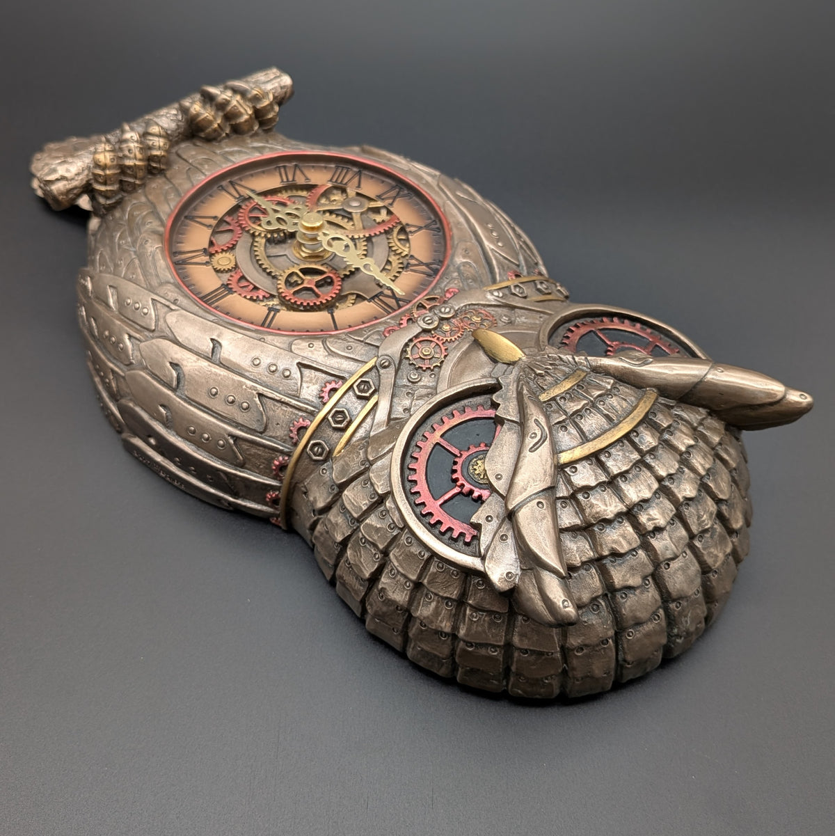 A steampunk-style decorative clock shaped like an owl, shown on a dark gray background. The object features textured metallic coloured surfaces in bronze, gold, and silver tones, with visible gears, and a circular clock face.