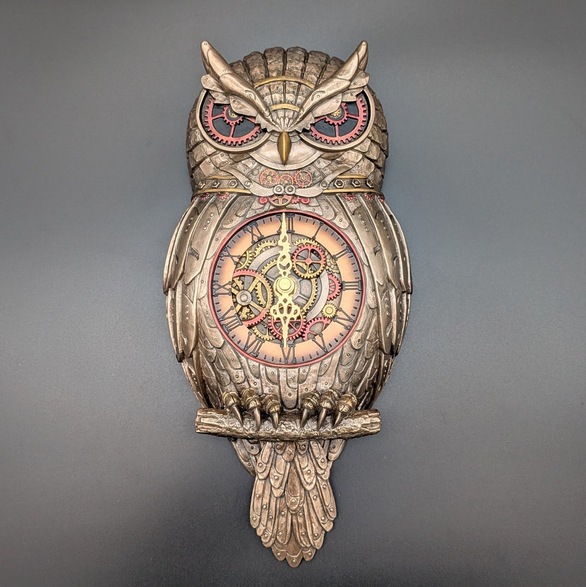 An ornate owl-shaped wall clock with a metallic coloured body, red and gold accents, and exposed clockwork details on the face, shown against a dark gray background.