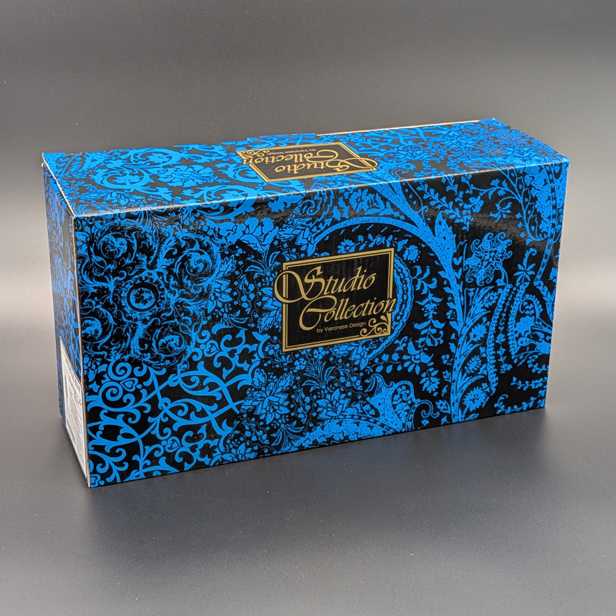 A rectangular product box with a blue and black ornate paisley pattern and gold 'Studio Collection' branding, photographed on a dark gray studio background.