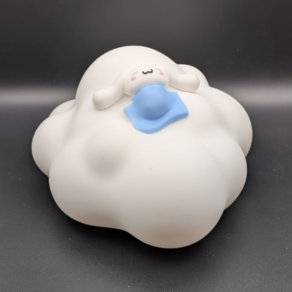 A white cloud-shaped decorative object with a small cartoon character peeking out from the top, under a blue blanket, set against a dark gray background.