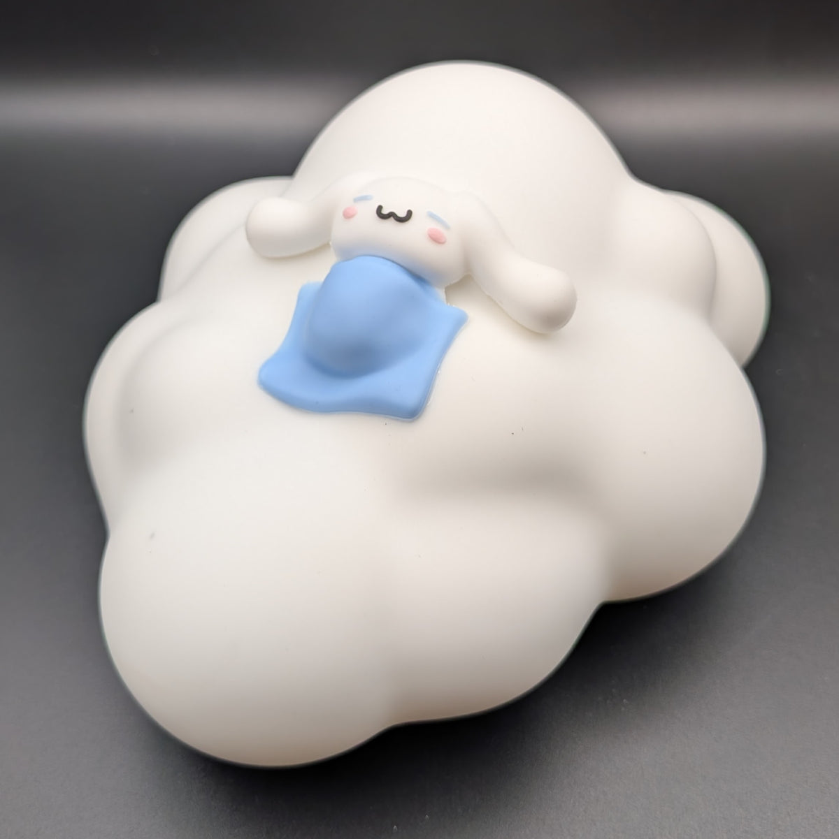 A white cloud-shaped decorative object with a small cartoon character peeking out from the top, holding a blue blanket, photographed on a dark gray background.
