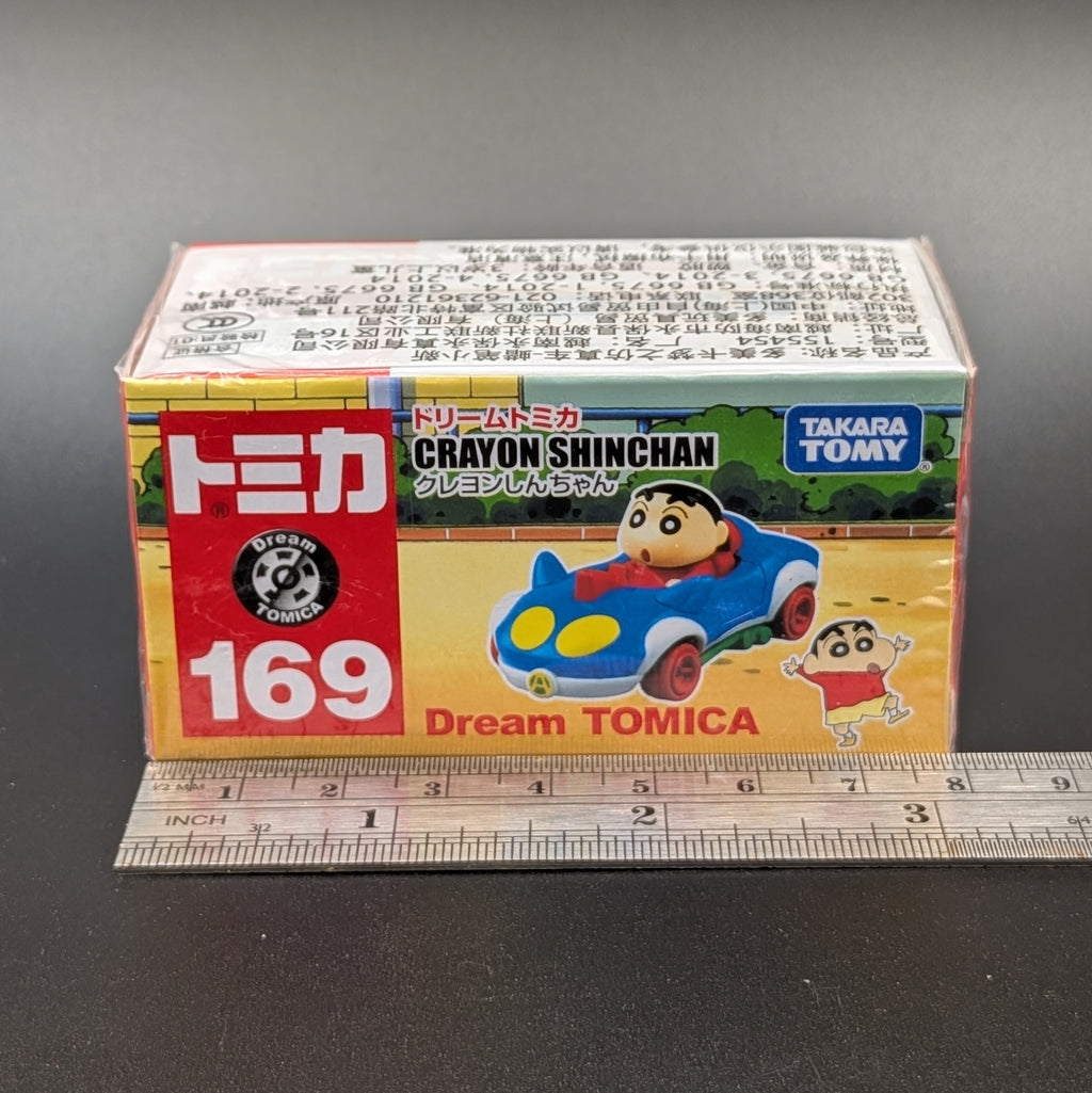 A boxed Tomica Crayon Shinchan model number 169, featuring the character Shinchan in a blue car, shown with a metal ruler in front to indicate scale against a dark gray background.