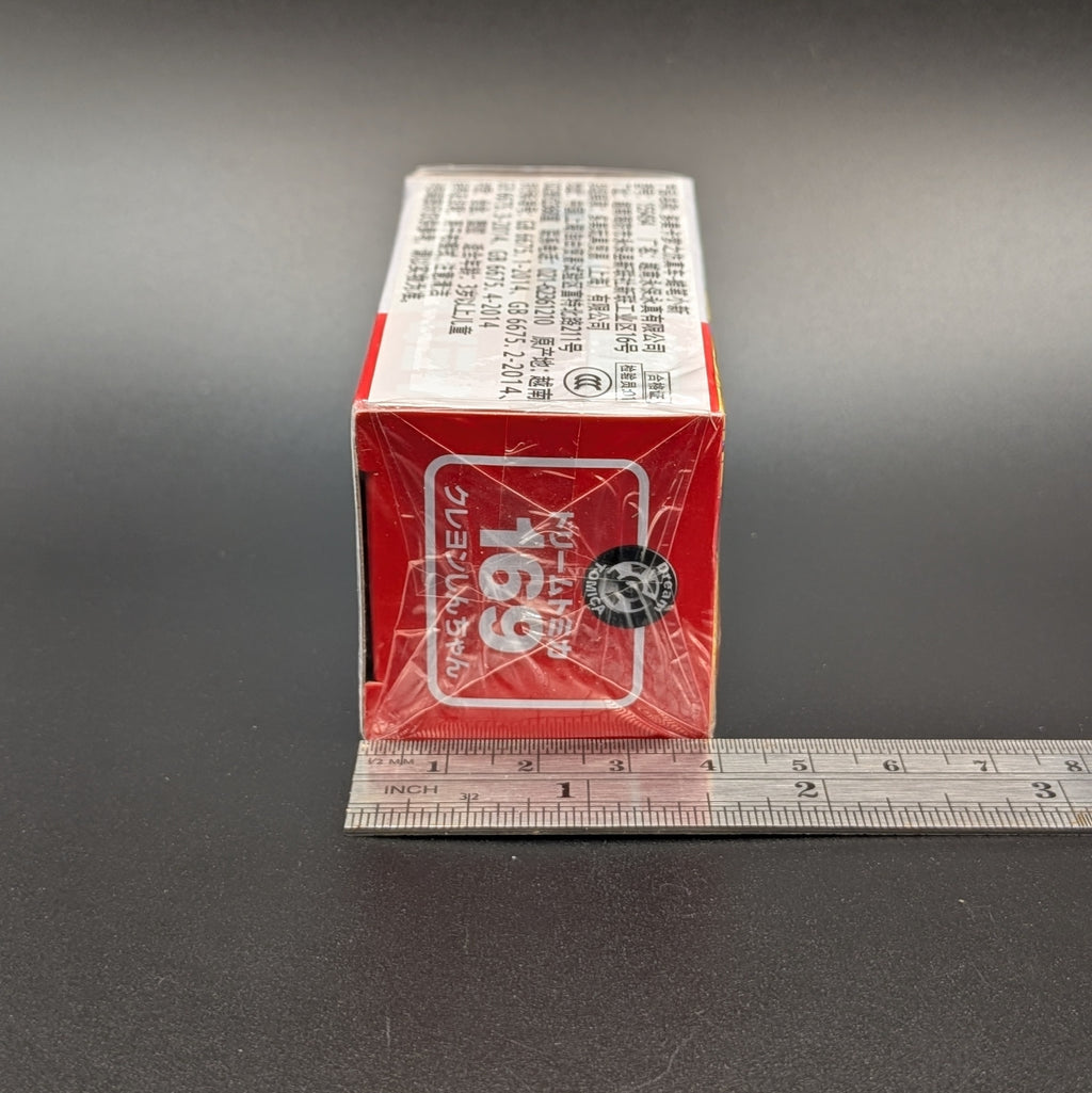 A red and white rectangular product box wrapped in clear plastic, shown  next to a metal ruler on a dark gray background.