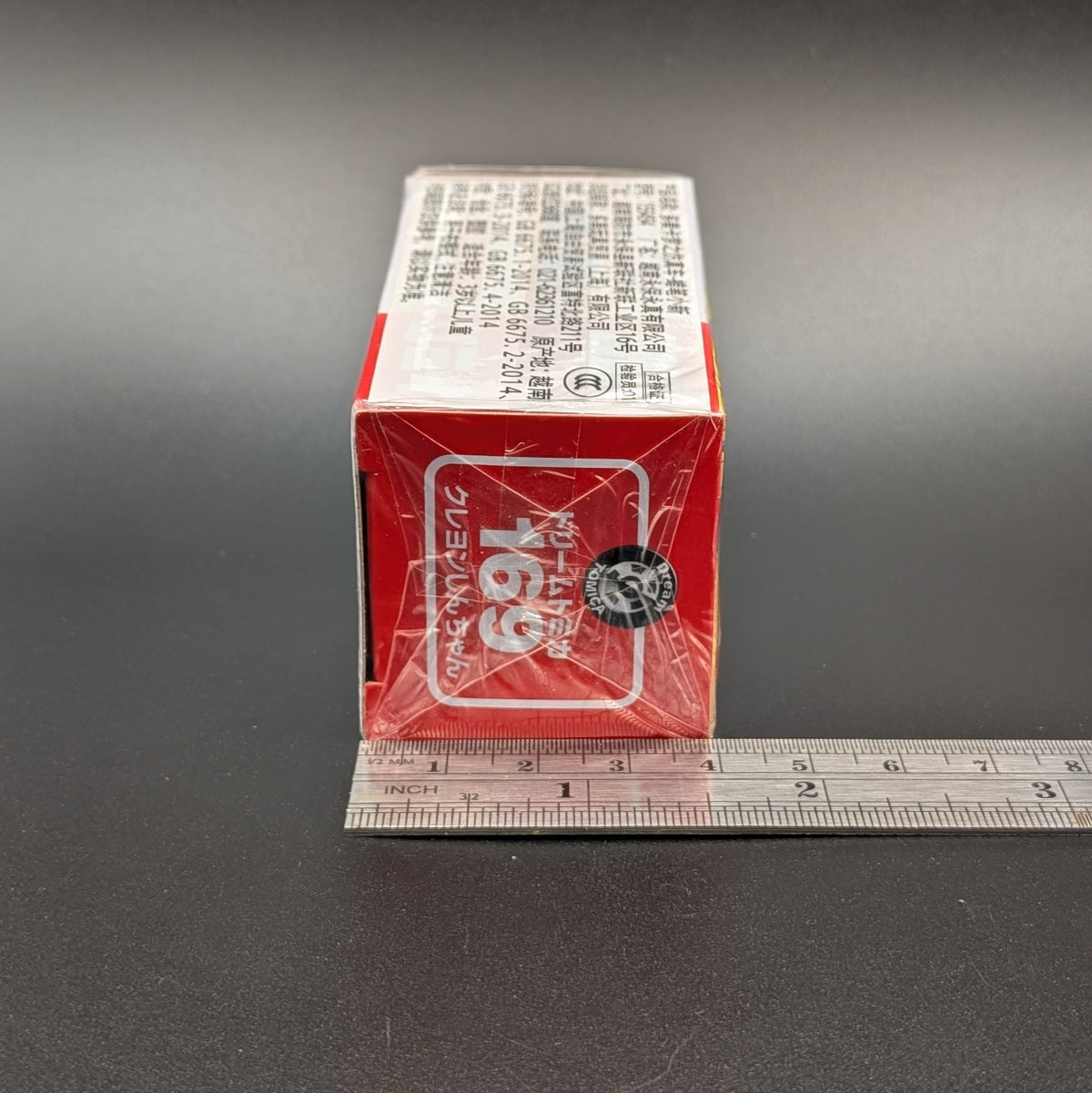 A red and white rectangular product box wrapped in clear plastic, shown  next to a metal ruler on a dark gray background.